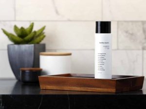Minimal bathroom scene with a white cylindrical shampoo bottle labeled “boldly basic” placed upright on a wooden tray on a dark countertop.
