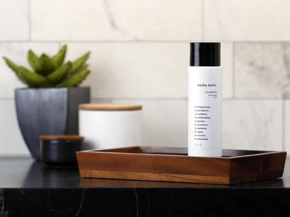 Minimal bathroom scene with a white cylindrical shampoo bottle labeled “boldly basic” placed upright on a wooden tray on a dark countertop.